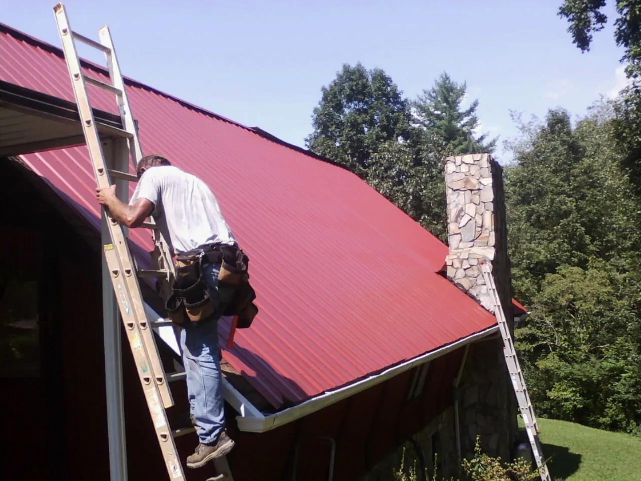 Roofer on a ladder beside a red metal roof and stone chimney — Soffit & Fascia Repair by Roofing Brothers in Bellaire