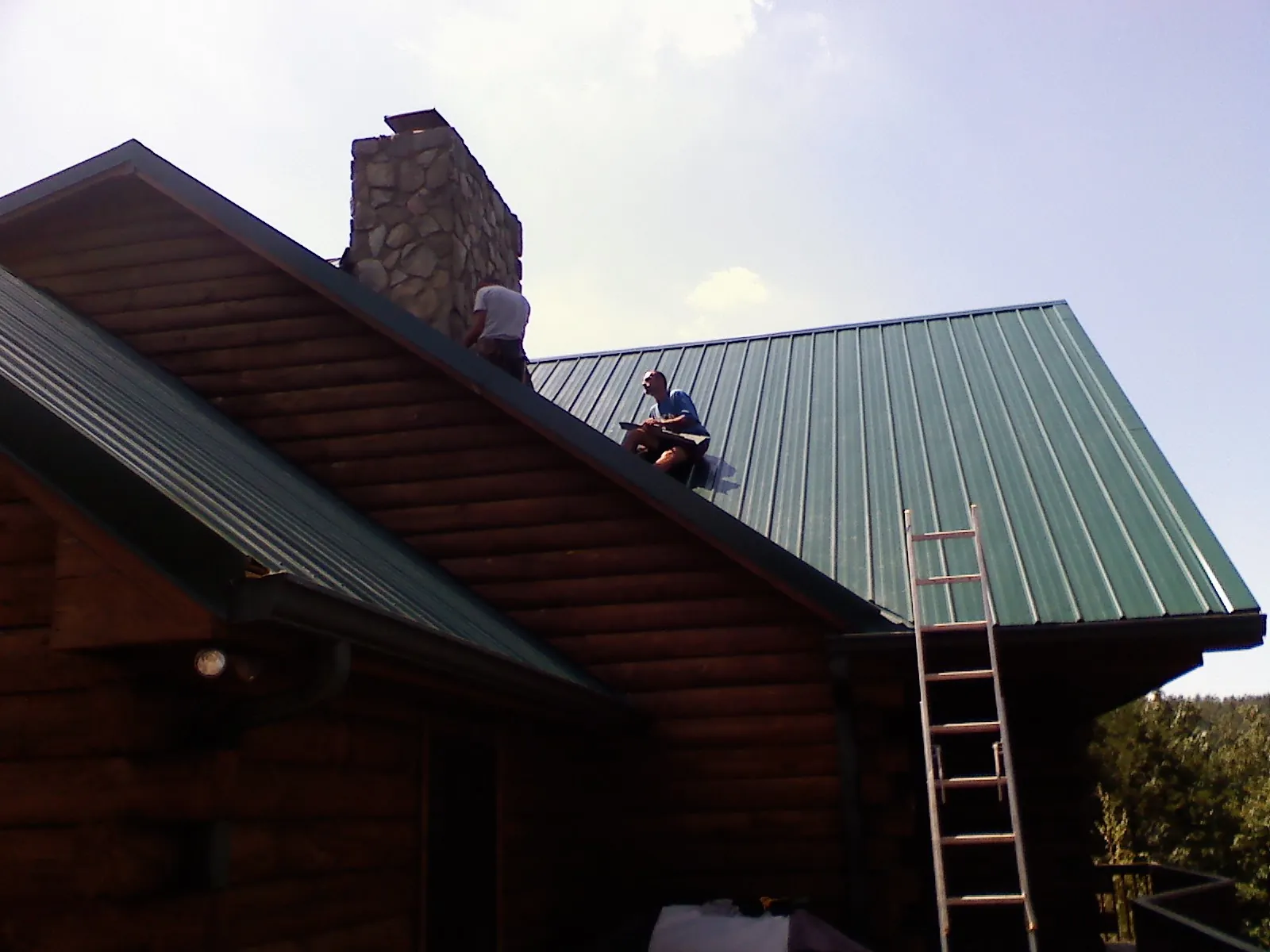 Roofers installing green metal panels beside a stone chimney on a log home in Bellaire